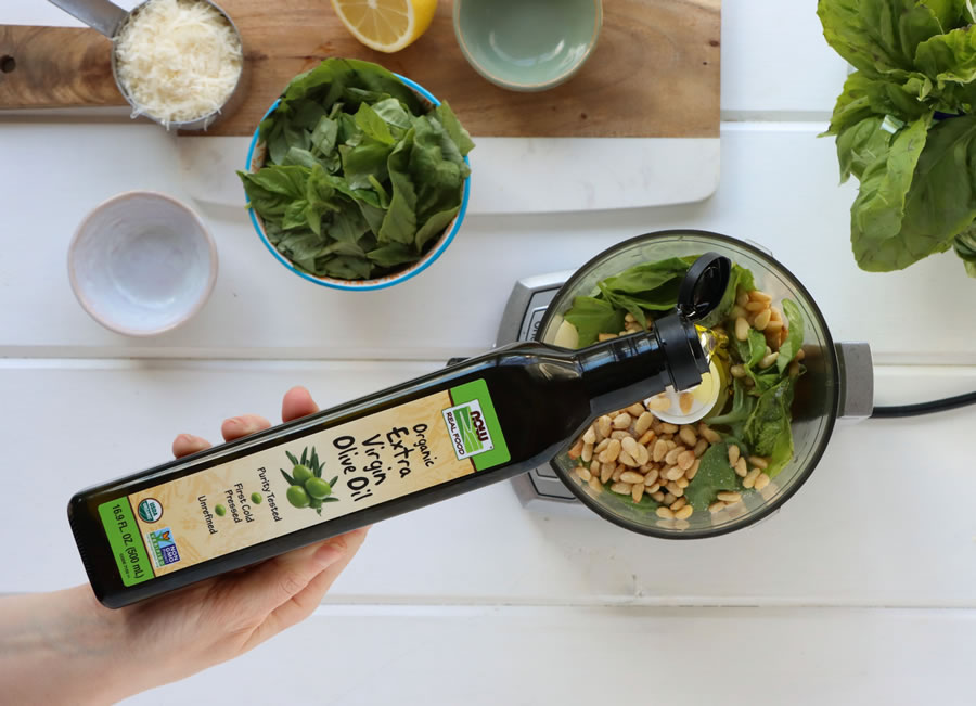 Now Foods Organic Extra Virgin Olive Oil being poured into a food processor with basil pesto ingredients