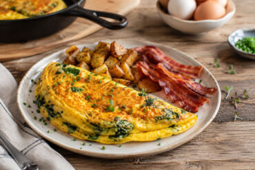 omelette with spinach and herbs served on a rustic ceramic plate with crispy bacon and roasted breakfast potatoes, on a wooden farmhouse table. a cozy luxury brunch atmosphere.