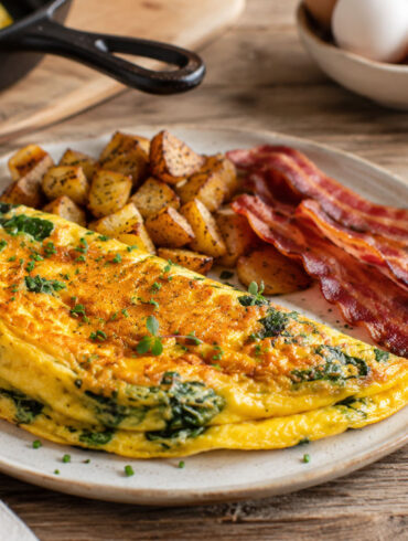 omelette with spinach and herbs served on a rustic ceramic plate with crispy bacon and roasted breakfast potatoes, on a wooden farmhouse table. a cozy luxury brunch atmosphere.