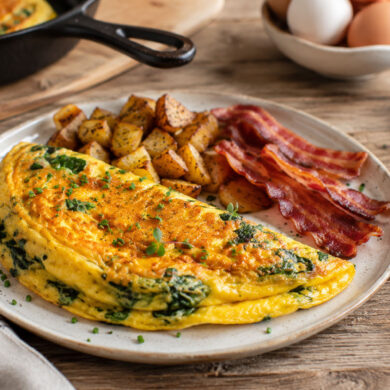 omelette with spinach and herbs served on a rustic ceramic plate with crispy bacon and roasted breakfast potatoes, on a wooden farmhouse table. a cozy luxury brunch atmosphere.