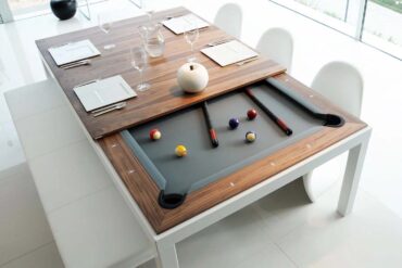 White and walnut Fusiontables dining pool table in modern dining room showing transformation from dining to pool table - wooden veneer top panels sliding to reveal gray felt playing surface with colorful billiard balls, pool cues, white contemporary chairs, wine glasses and place settings on remaining table top