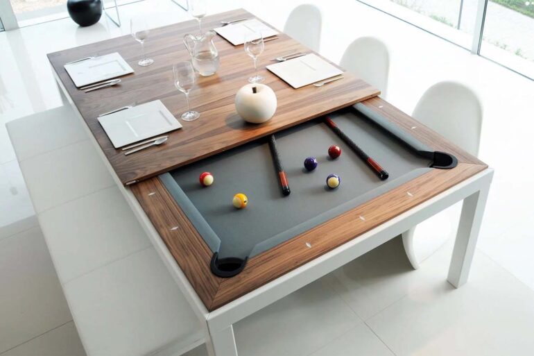 White and walnut Fusiontables dining pool table in modern dining room showing transformation from dining to pool table - wooden veneer top panels sliding to reveal gray felt playing surface with colorful billiard balls, pool cues, white contemporary chairs, wine glasses and place settings on remaining table top