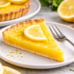 Slice of homemade lemon tart with bright yellow lemon filling and a buttery crust on a ceramic plate, topped with a fresh lemon slice and served with a fork, with whole lemons and another tart blurred in the background.