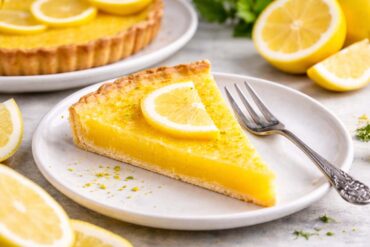 Slice of homemade lemon tart with bright yellow lemon filling and a buttery crust on a ceramic plate, topped with a fresh lemon slice and served with a fork, with whole lemons and another tart blurred in the background.
