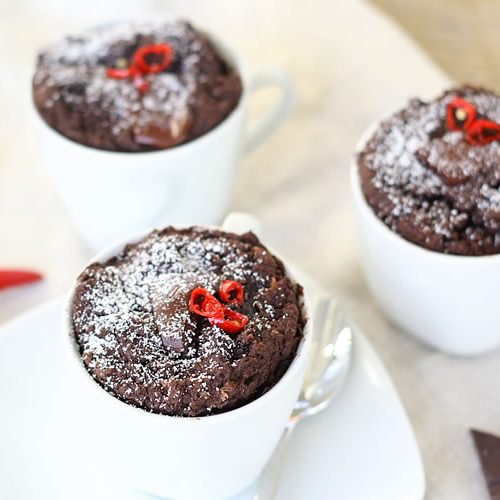 The image shows three chocolate mug cakes dusted with powdered sugar and garnished with small pieces of red chili peppers on top. The mug cakes are served in white coffee cups or mugs and appear to be freshly baked, with a rich, dark chocolate color. In the corner of the image, there are some pieces of chocolate bars and scattered red chili peppers, suggesting this is likely a spicy chocolate dessert that combines sweet and heat flavors. The cakes have a moist, cake-like texture and appear to have risen nicely in their mugs.