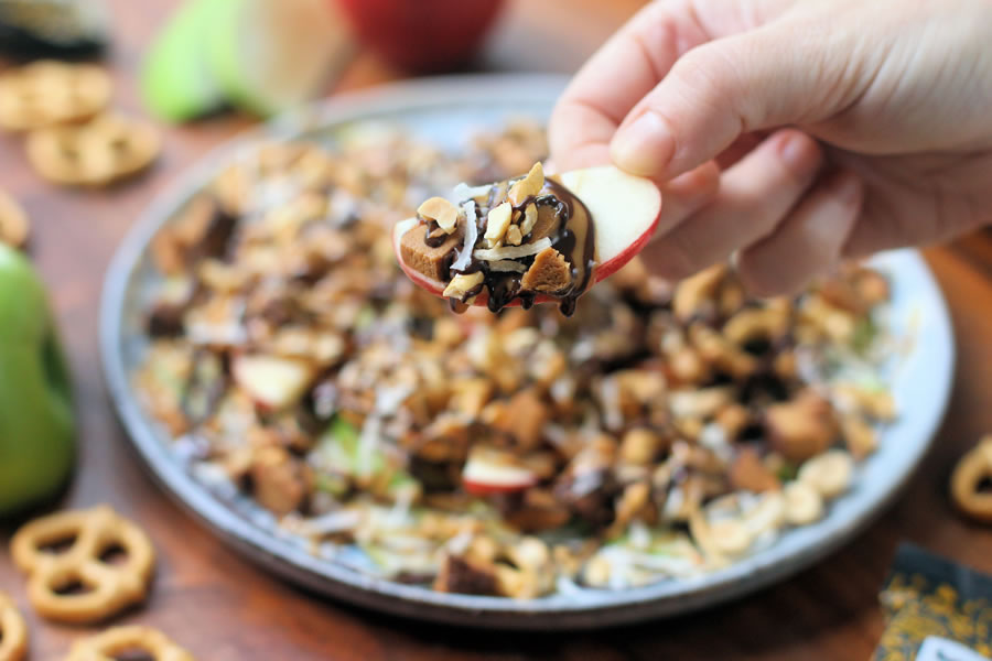 a hand holding an apple over a plate of apple nachos made with grain-free FitJoy pretzels melted chocolate peanut butter toasted coconut