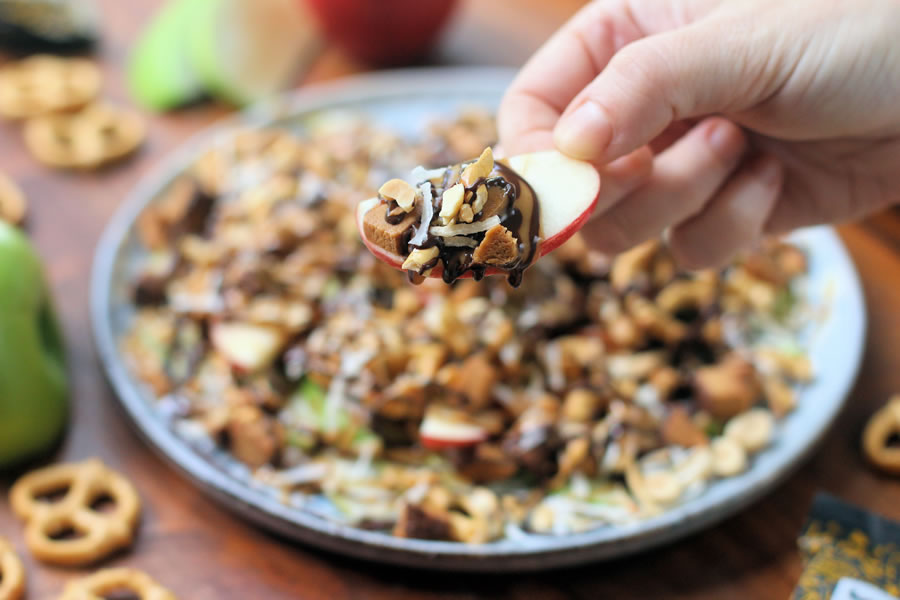 a hand holding up an apple slice with chocolate sauce peanut butter coconut peanuts and FitJoy protein bar crumble