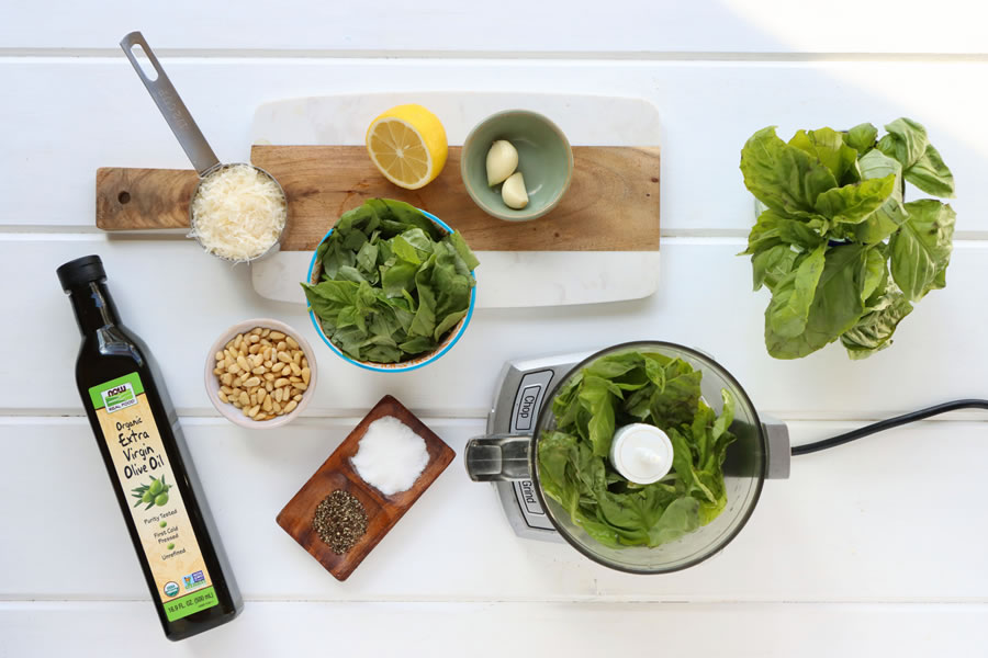 flatlay of ingredients laid out on a white table for basil pesto including fresh basil, pine nuts, lemon, garlic, parmesan cheese, extra virgin olive oil, salt and pepper