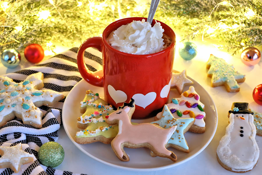 a plate of decorated sugar free christmas holiday cookies next to hot chocolate and christmast lights