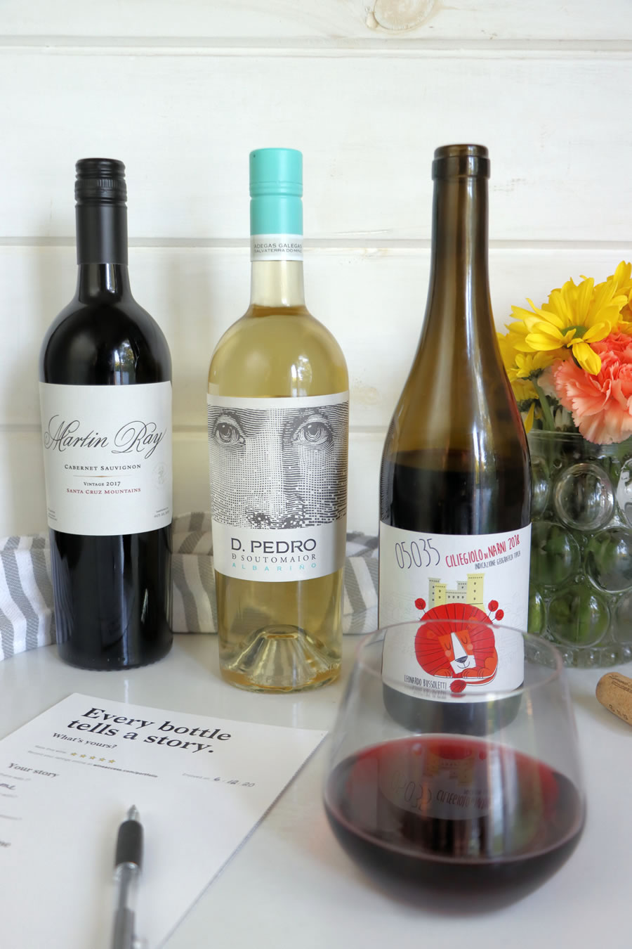 a selection of 3 wines from Wine Access wine club next to flowers a glass of red wine and a tasting card