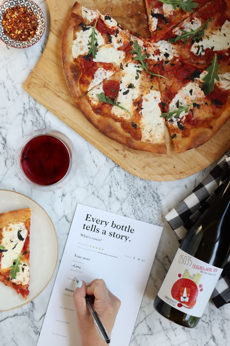 a hand filling out a wine tasting card and reviewing Wine Access next to pizza a glass of wine and a bottle