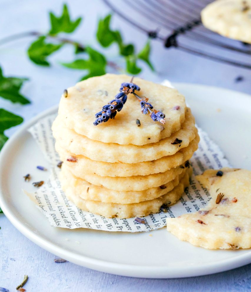 a stack of 6 buttery lavender shortbread cookies on a plate with lavender sprigs with lavender sprigs on newspaper