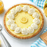 Greek yogurt lemon cream pie topped with whipped cream swirls and lemon slices in a graham cracker crust, styled on a bright white surface with fresh lemons and lemon curd.