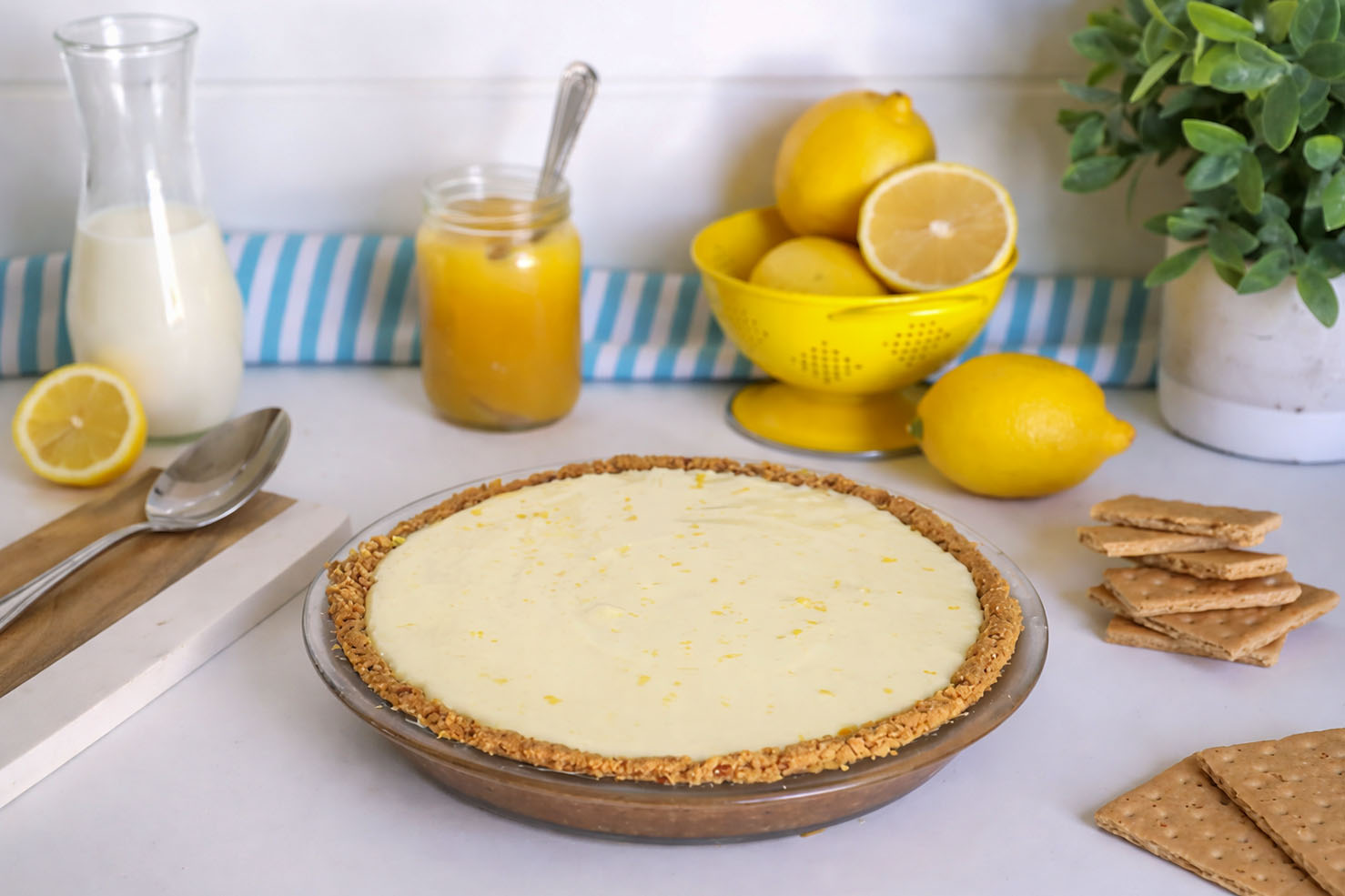 Unbaked Greek yogurt lemon cream pie in a graham cracker crust on a kitchen counter with lemon curd, fresh lemons, and graham crackers in natural light