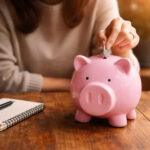 A woman at a table putting a coin into a piggy bank. Theres a notepad, pen, and coffee as if she's budgeting for a financial fast.