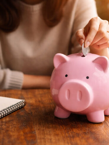 A woman at a table putting a coin into a piggy bank. Theres a notepad, pen, and coffee as if she's budgeting for a financial fast.