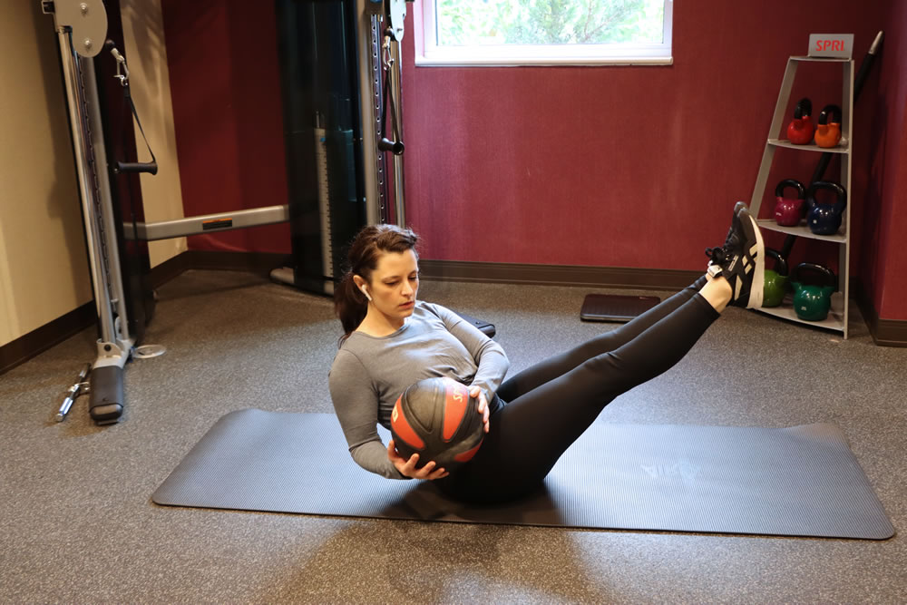 A woman lying on a yoga mat in a gym working out with a medicine ball.