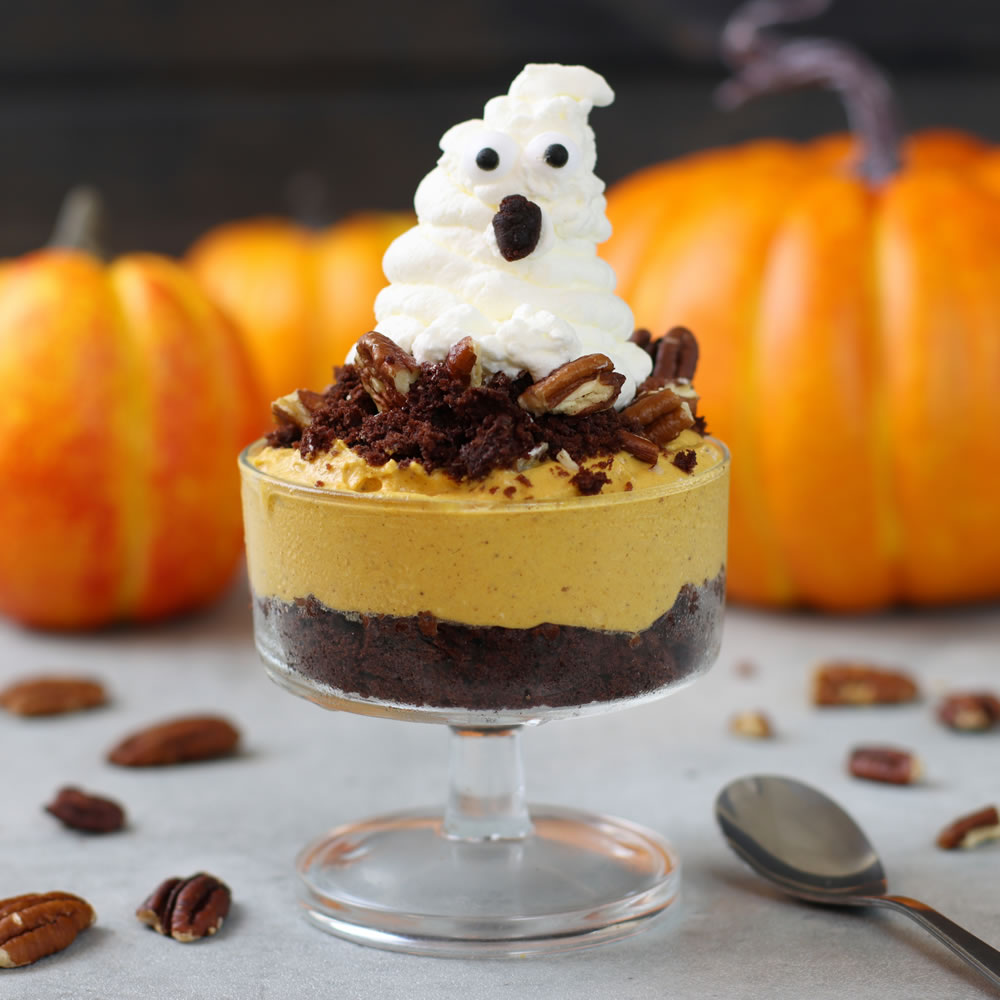 A close-up of a single glass dessert cup with layers of chocolate crumble, orange-colored mousse, and a whipped cream ghost on top. The ghost has chocolate chip eyes and mouth. Pecans are sprinkled on the mousse, and pumpkins are visible in the background.