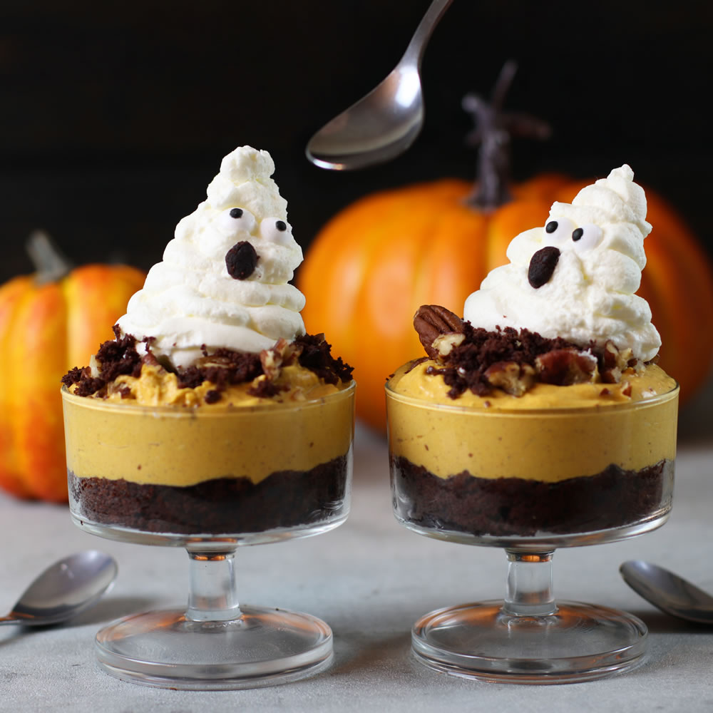 Two glass dessert cups filled with layers of chocolate crumble, orange-colored mousse, and topped with whipped cream ghosts. The ghosts have chocolate chip eyes and mouths. Pumpkins and spoons are visible in the background.