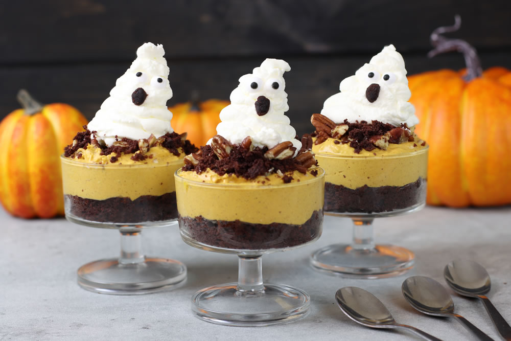 A set of clear dessert cups filled with layers of dark chocolate crumble, creamy pumpkin spice mousse, and topped with a ghost-shaped whipped cream dollop. The whipped cream ghosts have chocolate chip eyes and a small mouth made from chocolate, giving a cute, spooky appearance. Each cup is garnished with crumbled chocolate and pecans, and in the background, there are bright orange pumpkins that add to the festive Halloween theme.