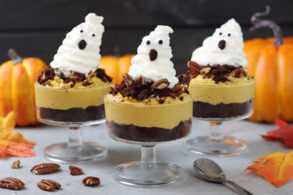 A set of clear dessert cups filled with layers of dark chocolate crumble, creamy pumpkin spice mousse, and topped with a ghost-shaped whipped cream dollop. The whipped cream ghosts have chocolate chip eyes and a small mouth made from chocolate, giving a cute, spooky appearance. Each cup is garnished with crumbled chocolate and pecans, and in the background, there are bright orange pumpkins that add to the festive Halloween theme.