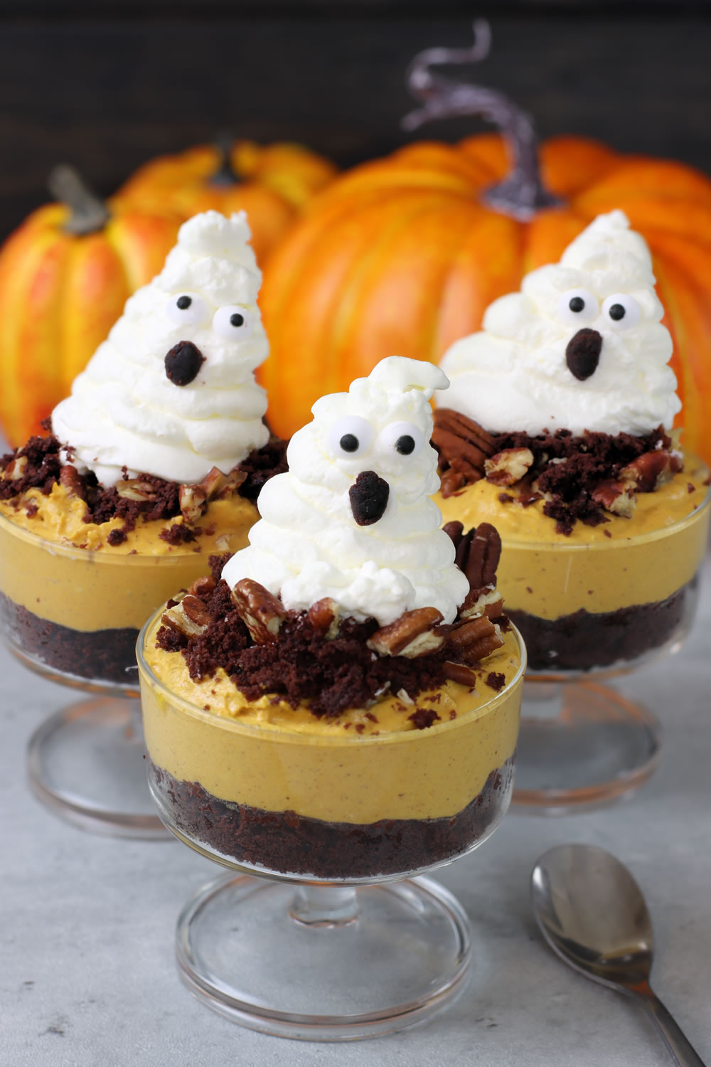 Three glass dessert cups filled with layers of chocolate crumble, orange-colored mousse, and topped with whipped cream ghosts. The ghosts have chocolate chip eyes and mouths. Pecans are sprinkled on the mousse. Pumpkins are visible in the background, and spoons are placed nearby.