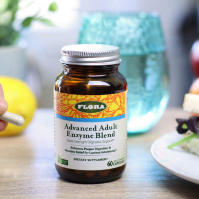 A woman hold a capsule of Advanced Adults Enzymes from Flora next to a sandwich and a glass of water.