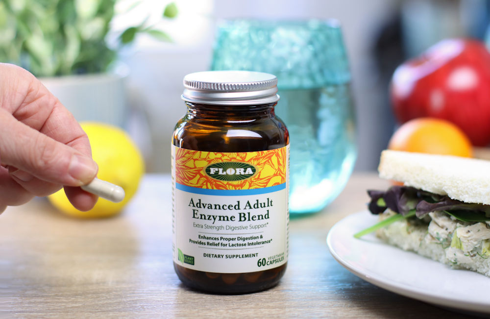 A woman hold a capsule of Advanced Adults Enzymes from Flora next to a sandwich and a glass of water.