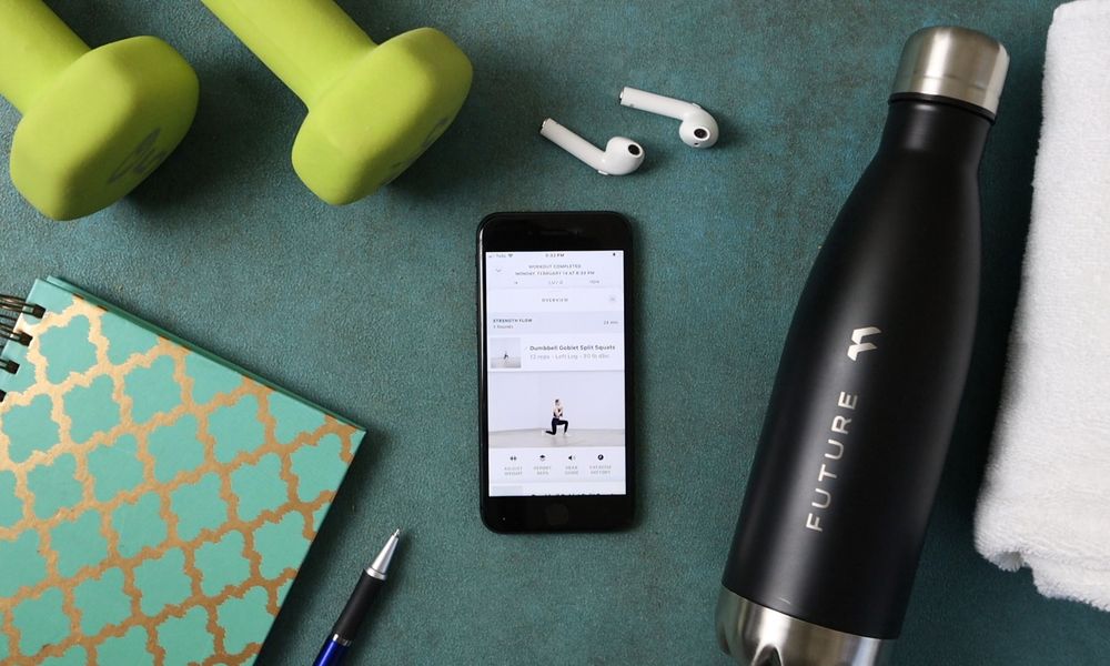 A dumbbell, water bottle, notebook, pen, earbuds, and iphone with the Future fitness app on a yoga mat.