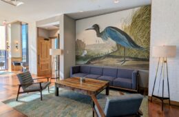 A sitting area to relax at The Rookery at the Merriweather Lakehouse Hotel in Columbia Maryland. A couch, table, two chairs, and a wall mural of a heron.