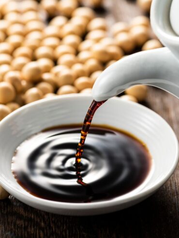 A bottle of soy sauce being poured into a bowl surrounded by soy beans.