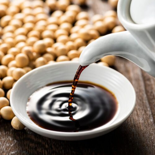 A bottle of soy sauce being poured into a bowl surrounded by soy beans.