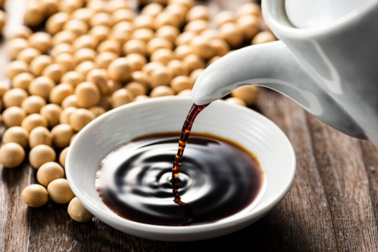A bottle of soy sauce being poured into a bowl surrounded by soy beans.