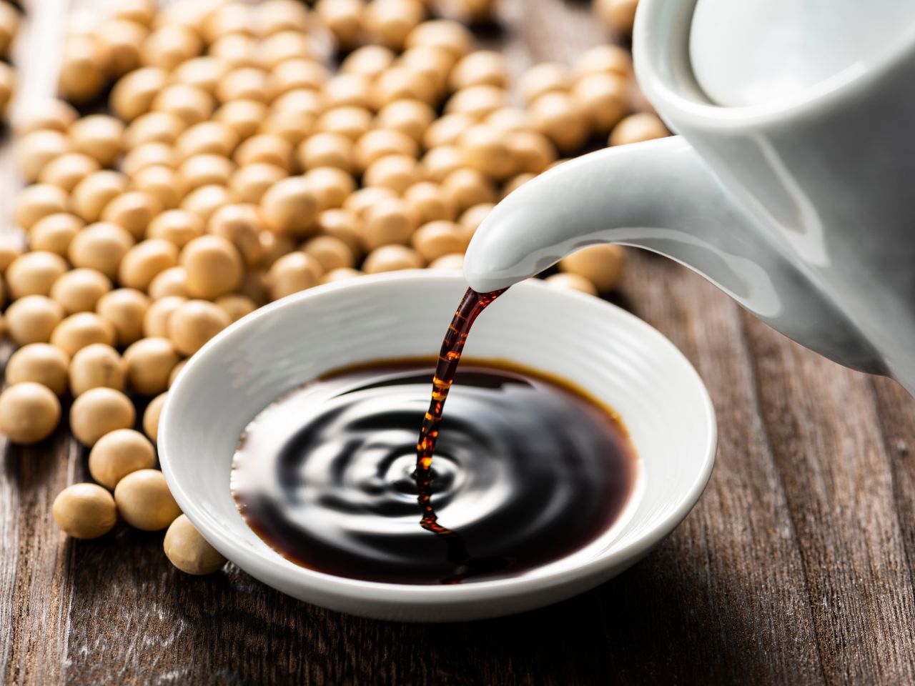A bottle of soy sauce being poured into a bowl surrounded by soy beans.
