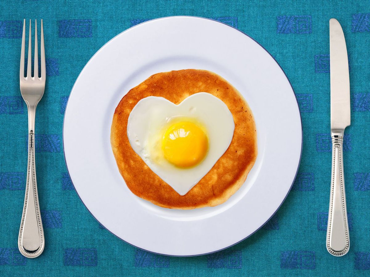 A heart shaped egg on a pancake on a plate next to a knife and fork.