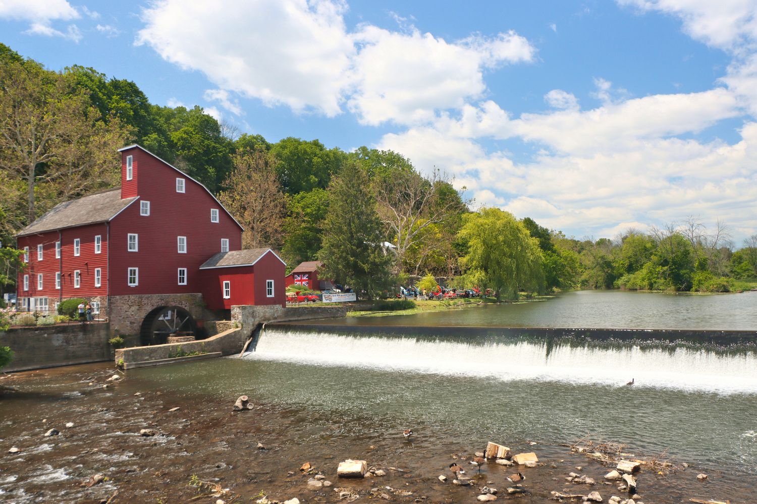 A picturesque historic red mill building sits alongside a small waterfall on the Raritan River in Clinton, New Jersey. The multi-story wooden structure features white-trimmed windows and a stone foundation. A small red outbuilding with a Union Jack flag is visible nearby. The mill is surrounded by lush green trees, and the scene includes a peaceful millpond with a dam creating a gentle waterfall. The bright blue sky with white clouds completes this quintessential American historical landscape.