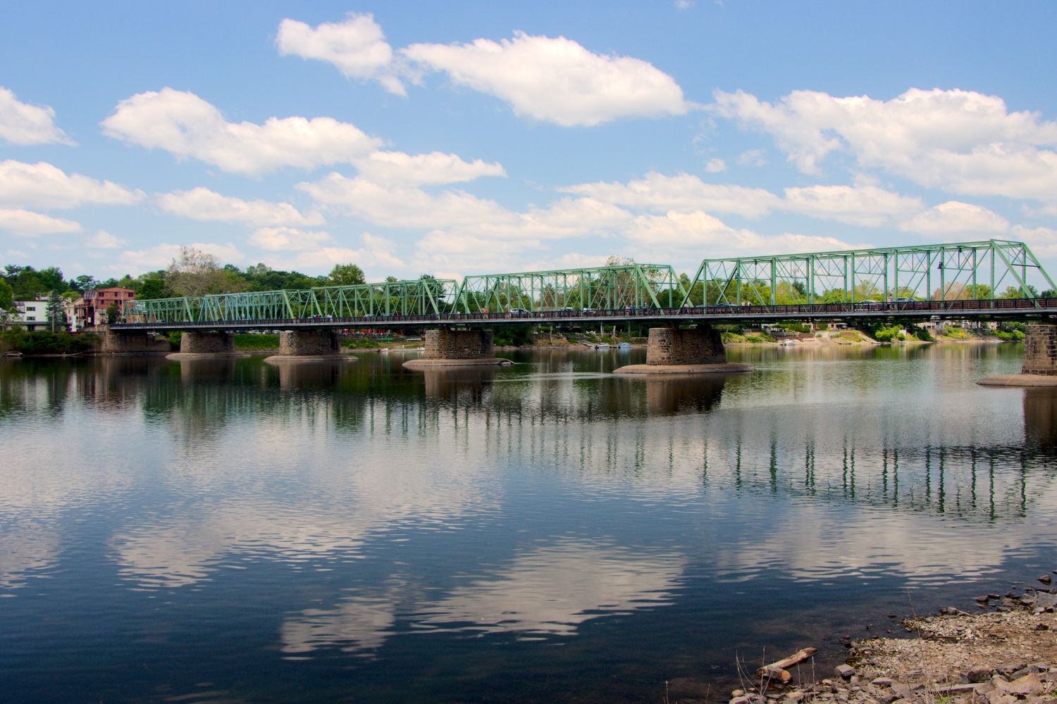 The historic green steel truss bridge that connects New Hope, Pennsylvania to Lambertville, New Jersey spanning across the Delaware River. The distinctive green-painted metal structure features multiple truss segments supported by stone pillars rising from the calm river below. The blue sky with scattered white clouds is perfectly reflected in the still water, creating mirror images of both the bridge and clouds. Buildings are visible on the shoreline, and the surrounding landscape shows lush spring or summer vegetation. This bridge serves as both a functional connection between the twin towns and a scenic landmark in the region.