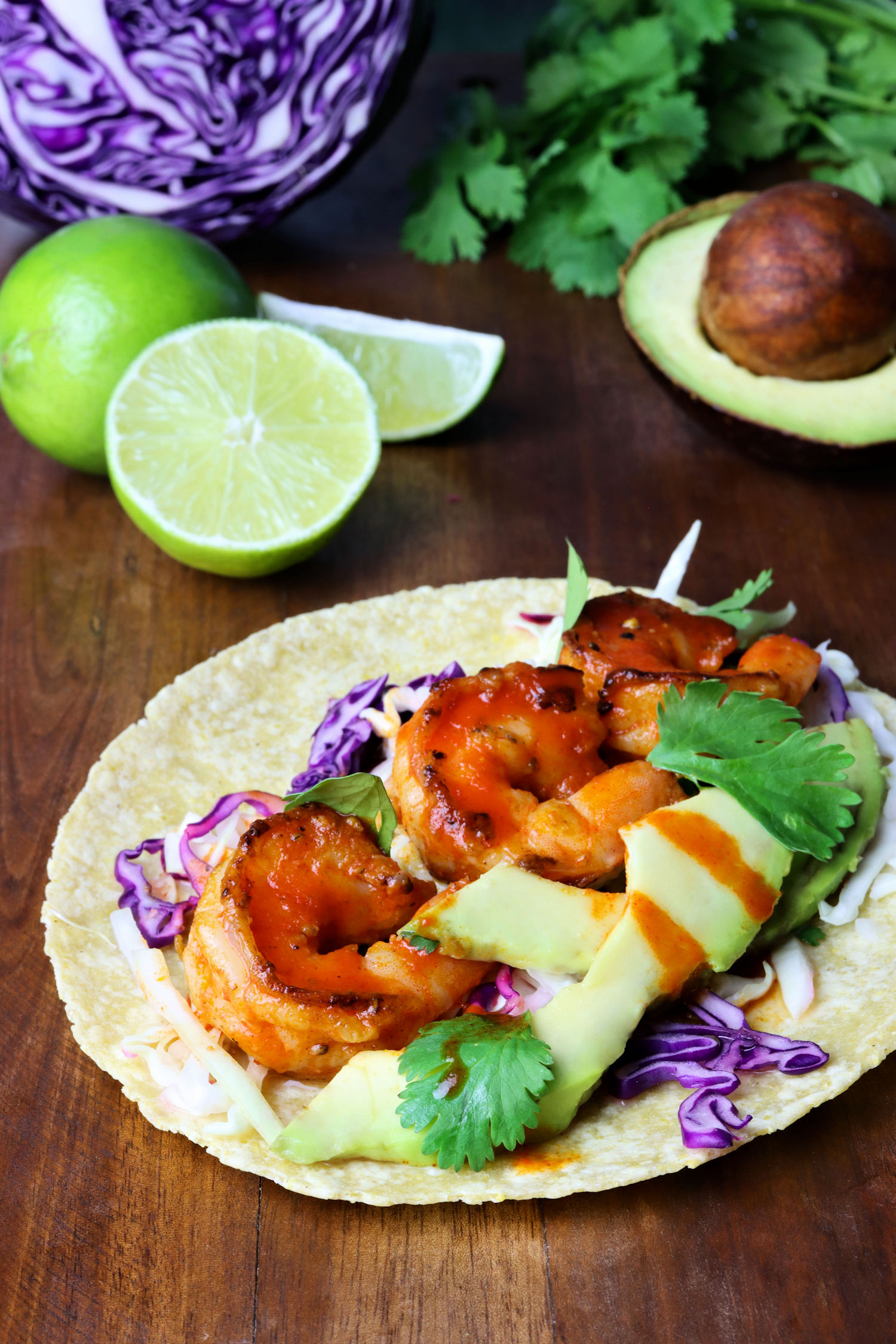 the completed recipe of spicy honey sriracha shrimp tacos on a table with ingredients, including lime, cilantro, red cabbage, and avocado