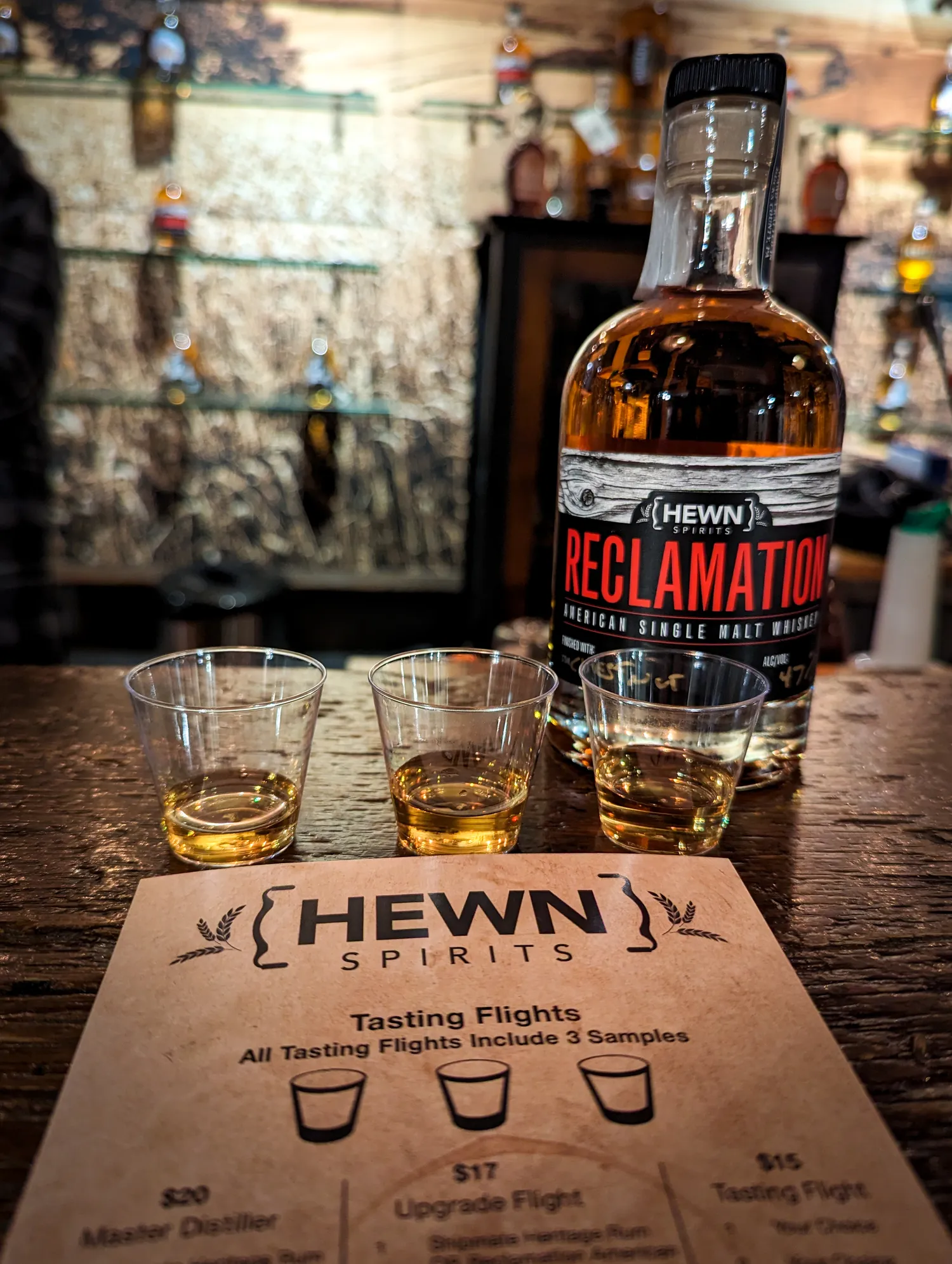 A bottle of Hewn Reclamation whiskey and 3 shots glasses lined up for a tasting.