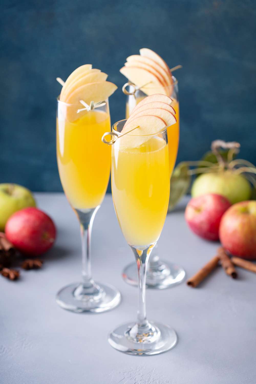 3 Apple Cider Mimosas in champagne glasses garnished with apple slivers with apples and cinnamon sticks in the background