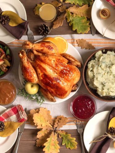 A traditional Thanksgiving dinner spread on a wooden table with fall decorations. The centerpiece is a golden-brown roasted turkey surrounded by sides including mashed potatoes, brussels sprouts, and a salad with apples and pecans. There's also pumpkin pie, cranberry sauce, and gravy. The table is decorated with pumpkins, autumn leaves, pinecones, and place settings with red and brown napkins. Several glasses of what appears to be apple cider are placed around the table.