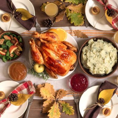 A traditional Thanksgiving dinner spread on a wooden table with fall decorations. The centerpiece is a golden-brown roasted turkey surrounded by sides including mashed potatoes, brussels sprouts, and a salad with apples and pecans. There's also pumpkin pie, cranberry sauce, and gravy. The table is decorated with pumpkins, autumn leaves, pinecones, and place settings with red and brown napkins. Several glasses of what appears to be apple cider are placed around the table.