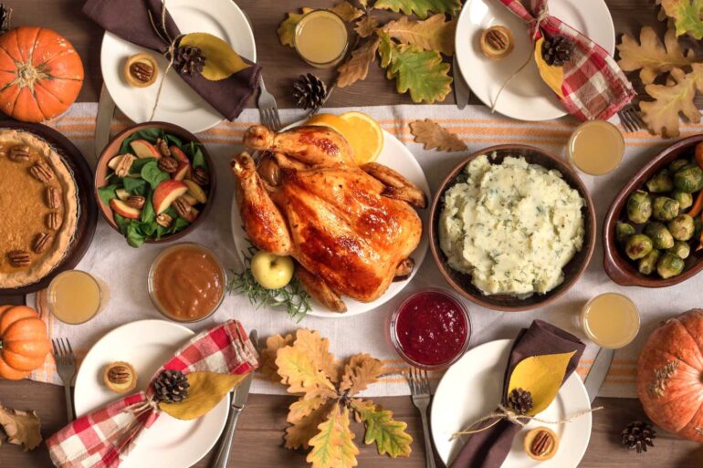A traditional Thanksgiving dinner spread on a wooden table with fall decorations. The centerpiece is a golden-brown roasted turkey surrounded by sides including mashed potatoes, brussels sprouts, and a salad with apples and pecans. There's also pumpkin pie, cranberry sauce, and gravy. The table is decorated with pumpkins, autumn leaves, pinecones, and place settings with red and brown napkins. Several glasses of what appears to be apple cider are placed around the table.