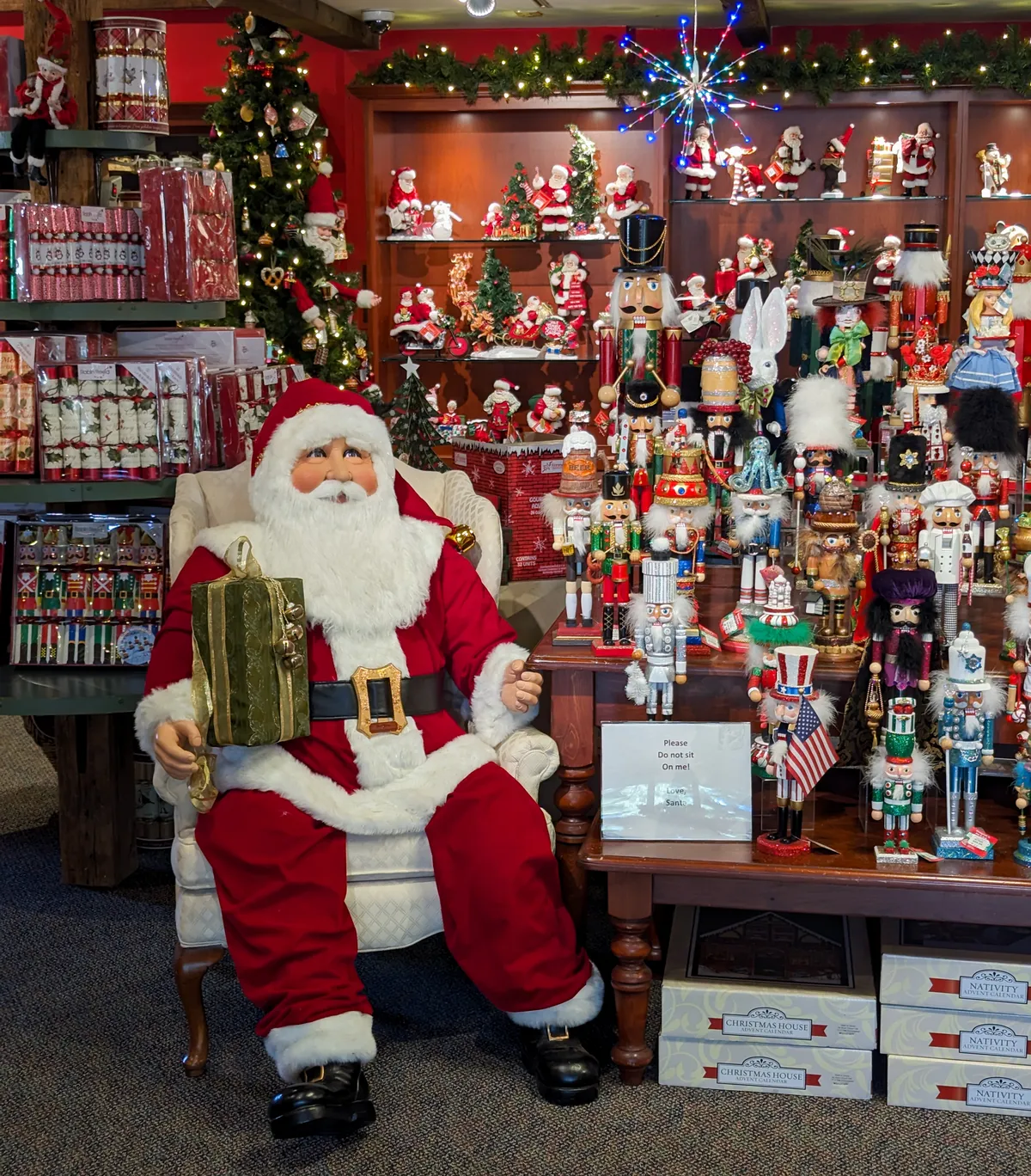 A Christmas display with a life size Santa sitting down next to a table of holiday decor. 