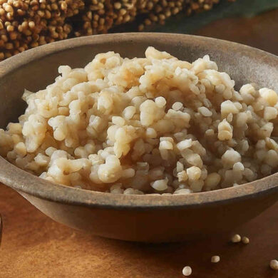 A rustic bowl of cooked sorghum on a table with uncooked sorghum and sorghum plants.