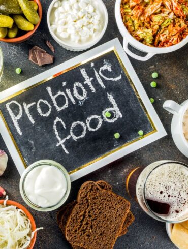 This image showcases a variety of probiotic foods arranged around a small chalkboard that says "Probiotic food." The assortment includes: Pickles in a small dish A bowl of cottage cheese A jar of sauerkraut Kimchi Glasses of yogurt and kefir A plate of soft cheese Dark chocolate squares Rye bread slices A bowl of peas A head of garlic Parmesan cheese pieces The overall theme emphasizes fermented foods and other natural sources of probiotics.