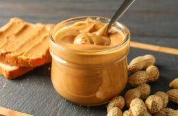 A jar of peanut butter with a spoon next to unshelled peanuts and peanut butter on bread.