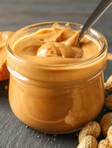 A jar of peanut butter with a spoon next to unshelled peanuts and peanut butter on bread.