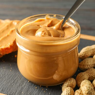 A jar of peanut butter with a spoon next to unshelled peanuts and peanut butter on bread.