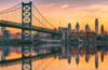 A romantic view of the Philadelphia skyline, water, and bridge at sunset.